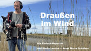 Ein Mann steht mit Mikrofon draußen im Schilf. Im Hintergrund ist ein Leuchtturm.